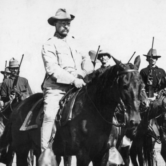 Theodore Roosevelt and the Rough Riders on horses with guns.