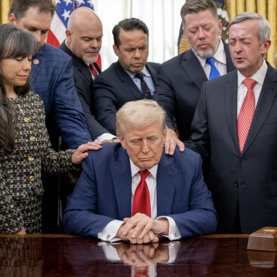 White people close their eyes in prayer while laying hands on Donald Trump at his desk in the Oval Office.