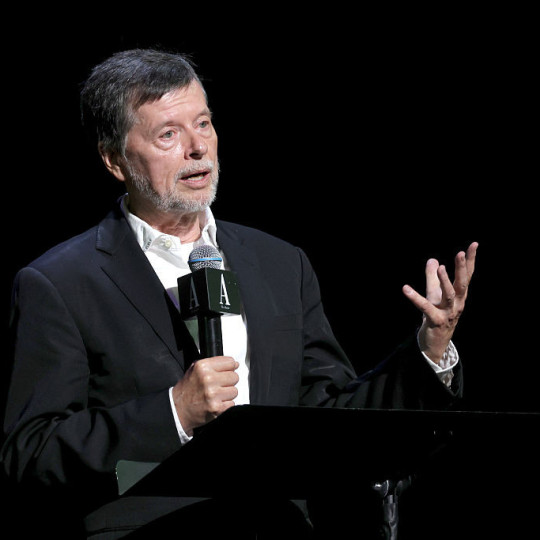 Ken Burns stands at a podium, one hand holding a microphone, the other gesturing as he presents.