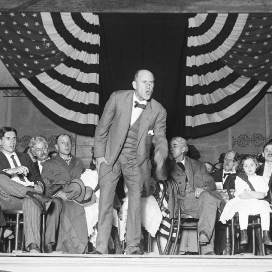 Eugene Debs passionately addresses a crowd from atop a stage, with American flags draped in the background.