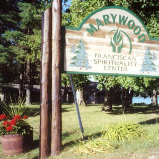 A rustic sign advertises Marywood Spirituality Center. Trees, flowers, and a view of the lake lie in the background.