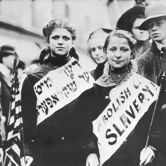 Jewish immigrant women protesting wearing banners in English and Yiddish and holding American flags.