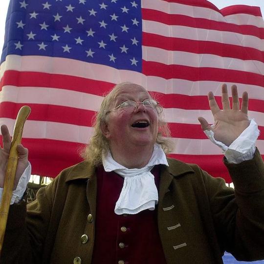 A Benjamin Franklin impersonator comedically raising his hand. An American flag waves in the background.