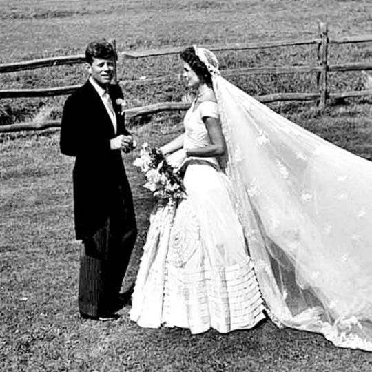 Jackie Kennedy stands in a flowing white gown with a white veil billowing out behind her.