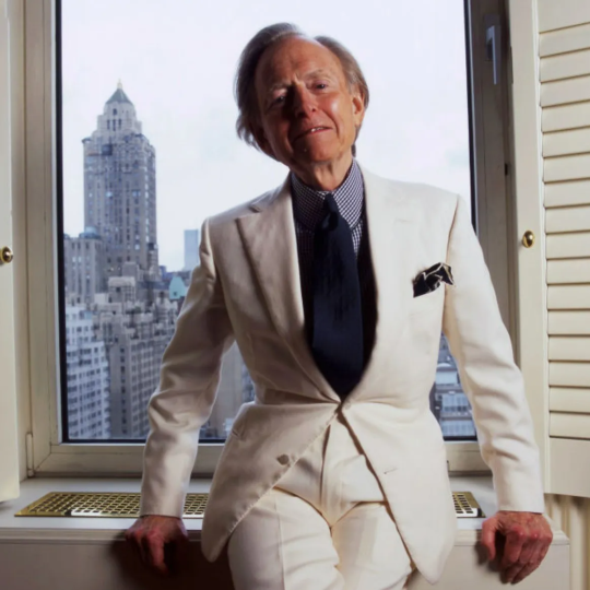 Tom Wolfe leans on the window sill in his classic white suit, the New York skyline in the background.
