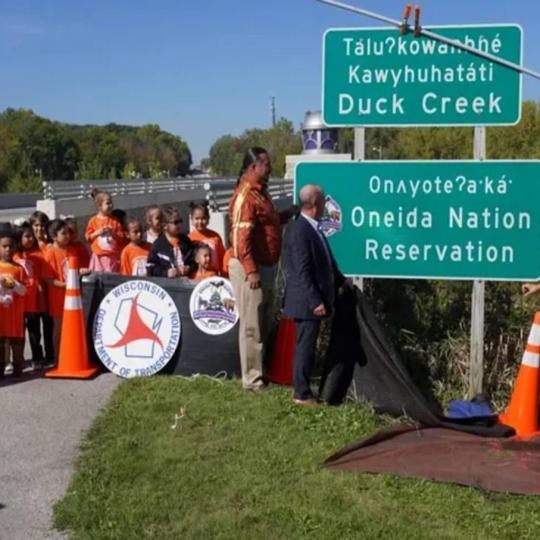 A group of children observe the unveiling of street signs in English and Oneida.