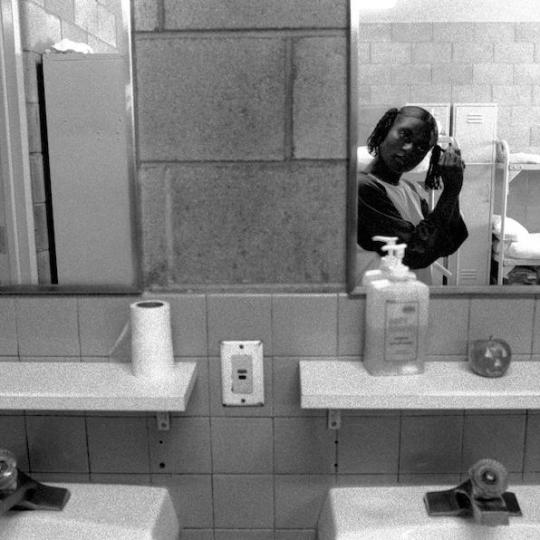 In a prison bathroom, a girl does her hair while a guard looks on.
