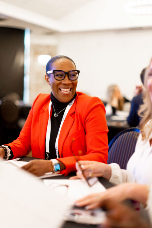 Person in a red blazer smiling and sitting at a table in a conference setting, holding a pen.