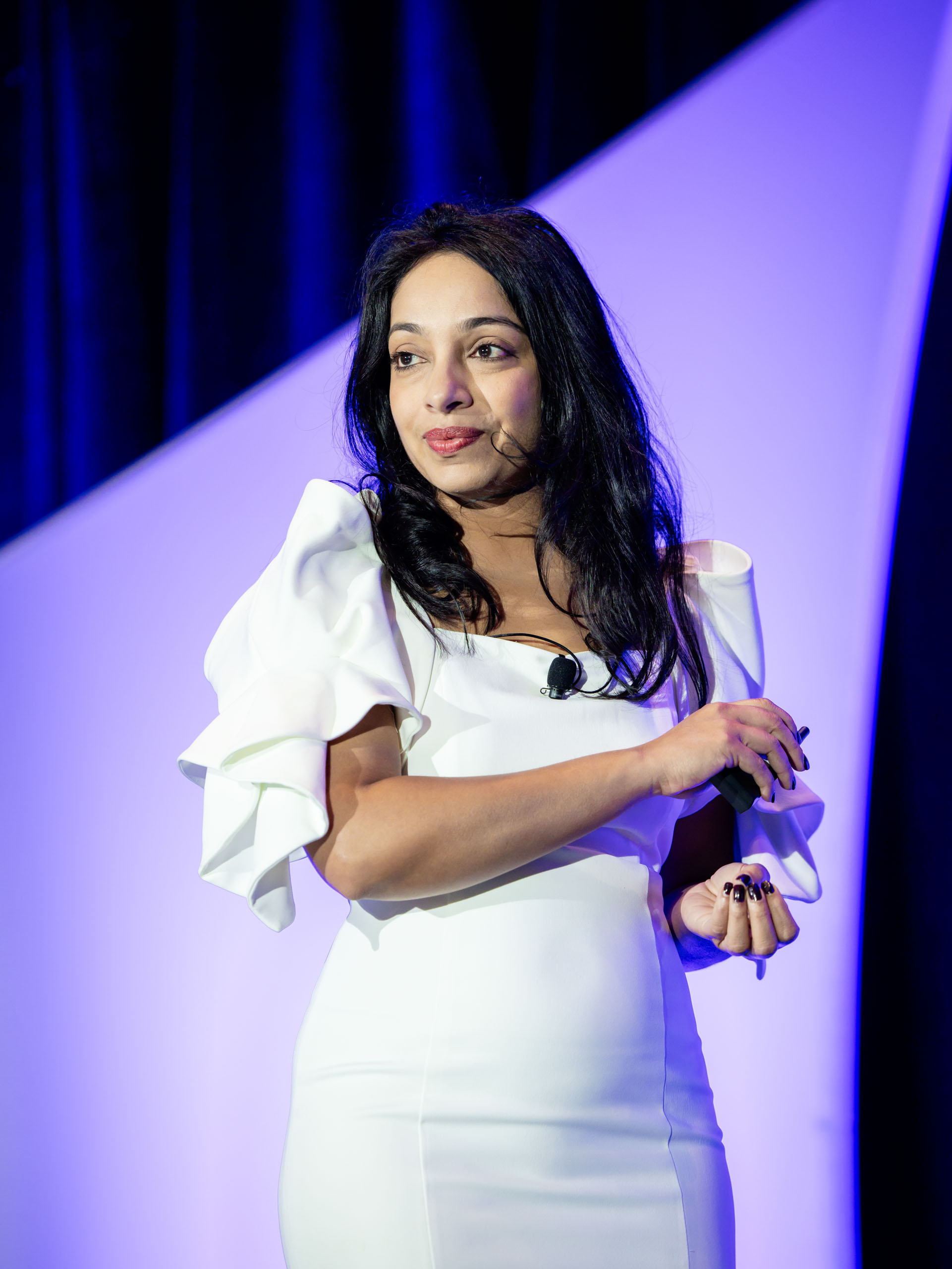 A woman in a white dress speaks on stage, holding a microphone. A purple backdrop is behind her.