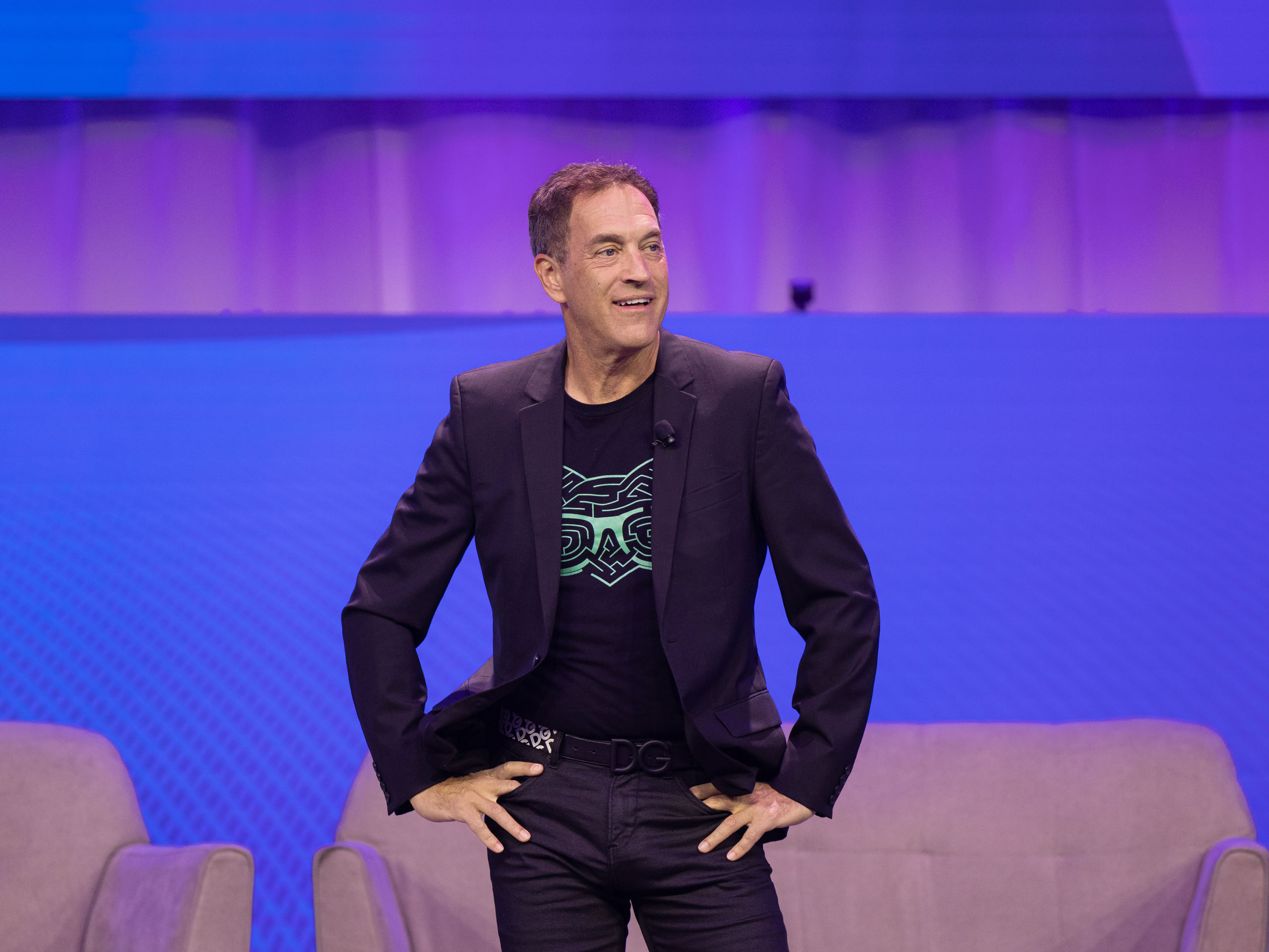 A man in a black suit and graphic t-shirt stands confidently on stage, with two beige chairs in the background against a blue backdrop.