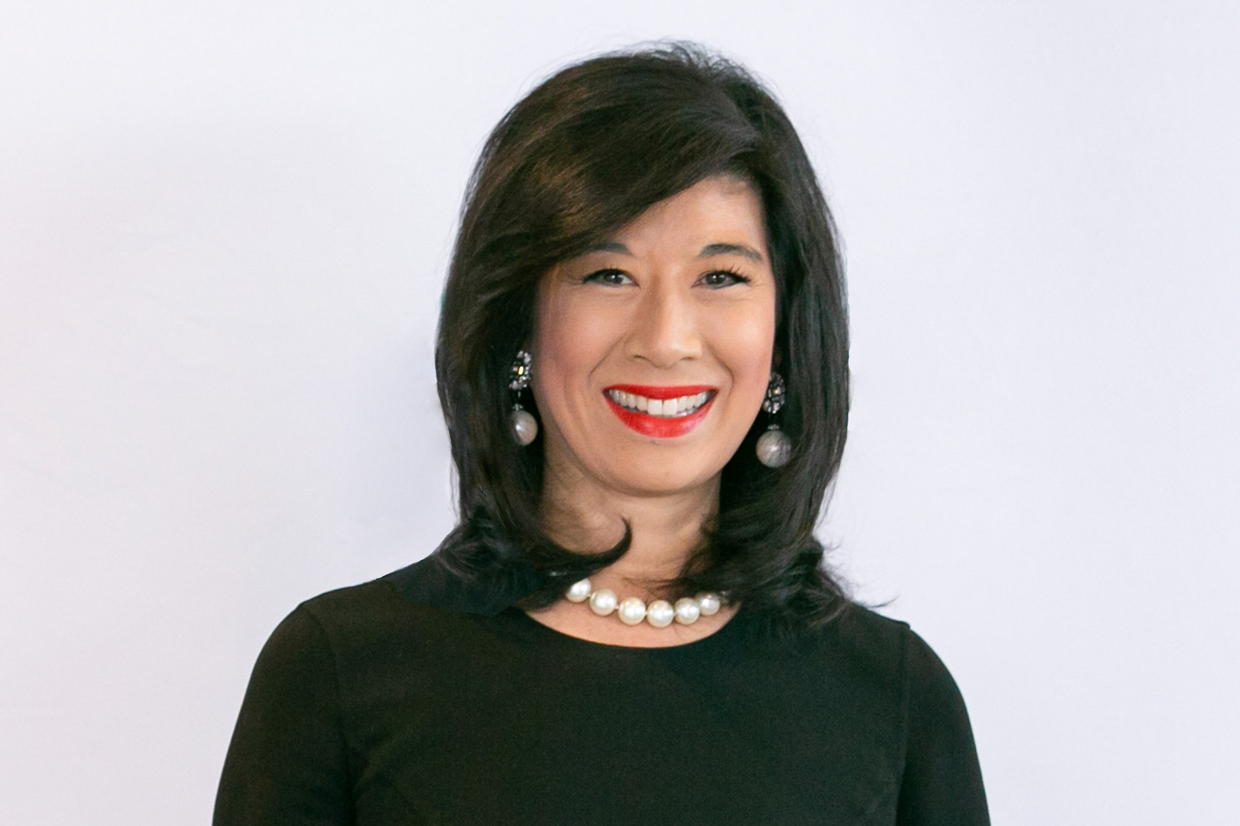 A person with shoulder-length dark hair, wearing a black top, pearl necklace, and earrings, smiles against a plain background.
