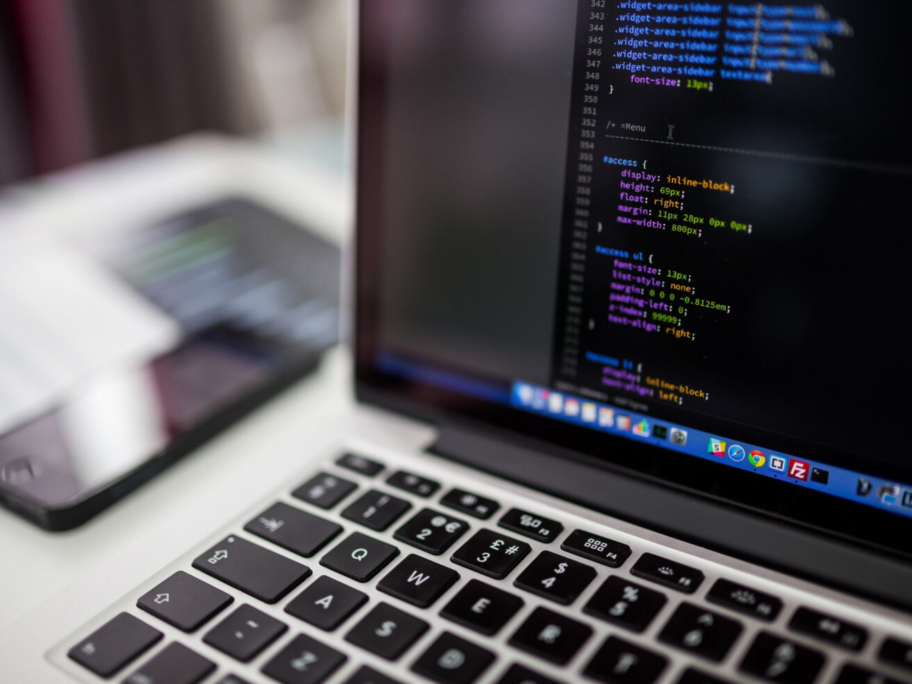 A laptop displaying code on the screen sits next to a smartphone on a desk.