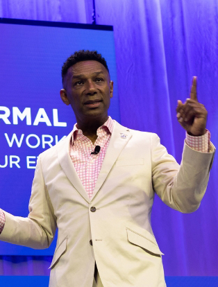 A man in a beige suit gestures while speaking on stage against a blue backdrop.