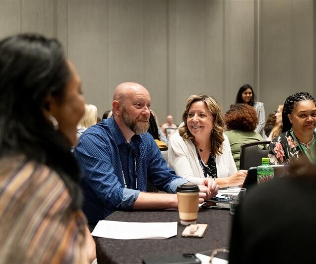 A group of people sit around a table in a conference room, engaged in conversation. Notebooks, a coffee cup, and a soda can are on the table.
