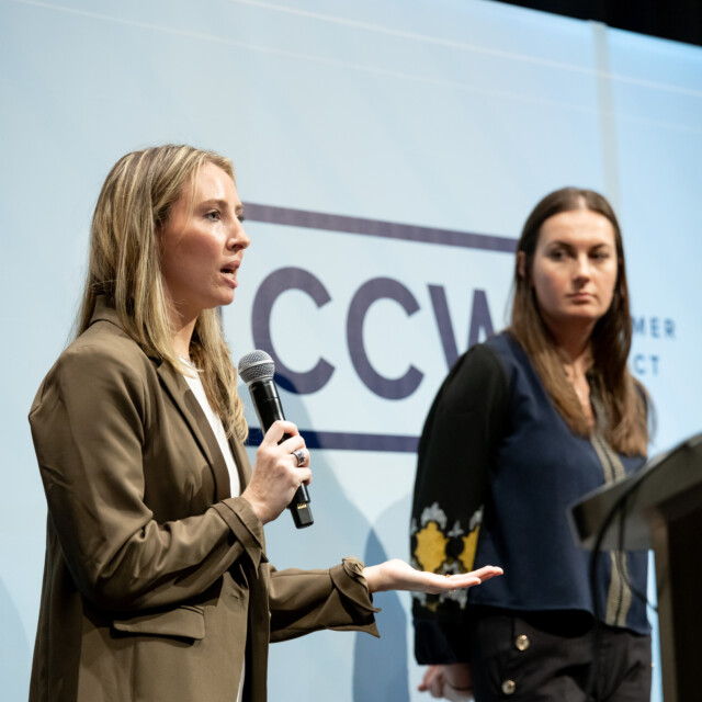 Two women stand on stage; one speaks into a microphone while the other listens near a podium with a laptop. A "CCW" sign is visible in the background.