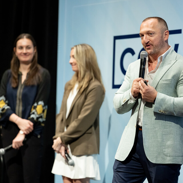 A man speaks into a microphone on stage while two women stand in the background near a sign that reads "Customer Contact.