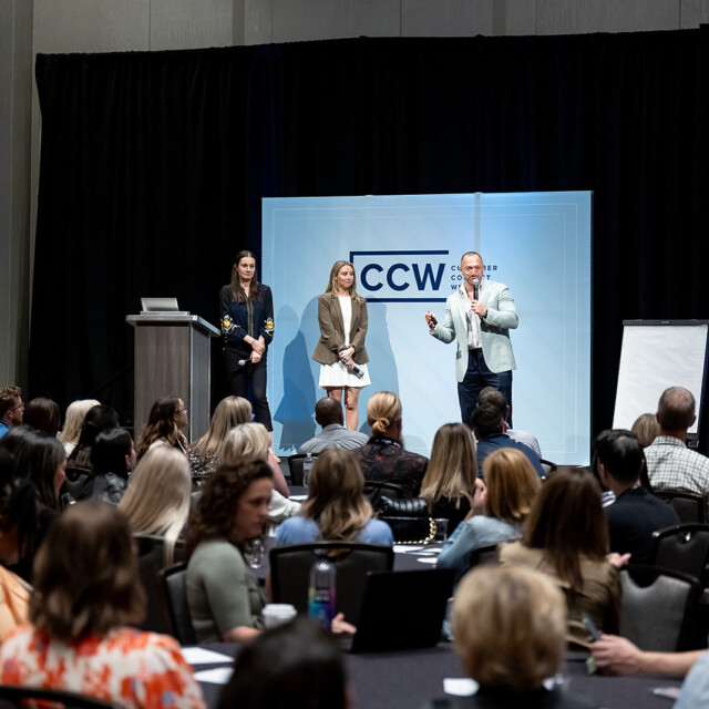 Three presenters stand on stage speaking to a seated audience at a conference, with a "CCW" sign and a blank easel in the background.