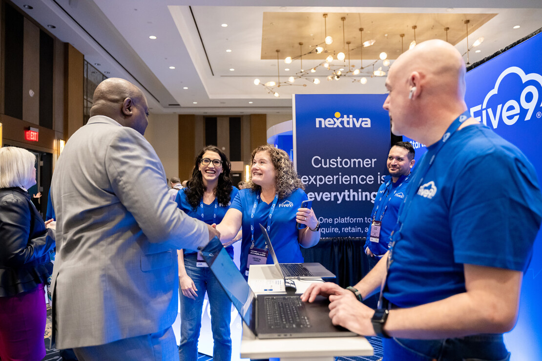 A group of people wearing blue "Five9" shirts interact with a visitor at a technology conference booth displaying the Nextiva and Five9 logos.