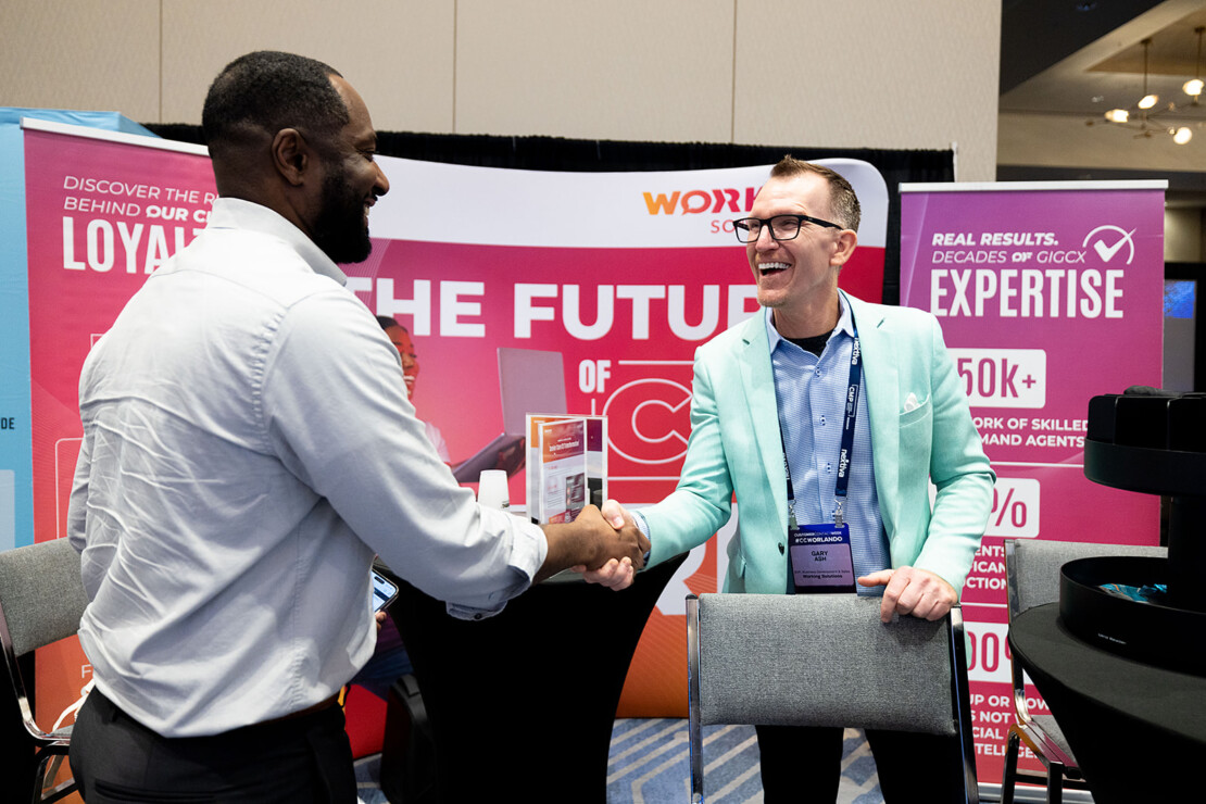 Two men shake hands and smile at a conference booth featuring banners with text about loyalty, expertise, and customer experience.