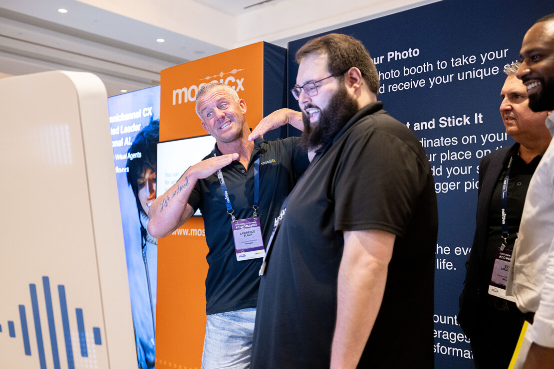 Four men stand in front of a photo booth at a conference; one man poses playfully while others smile and look at the booth screen.