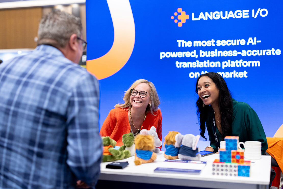 Two women smile and talk with a man at a conference booth for Language I/O, with plush toys and promotional items on the table.