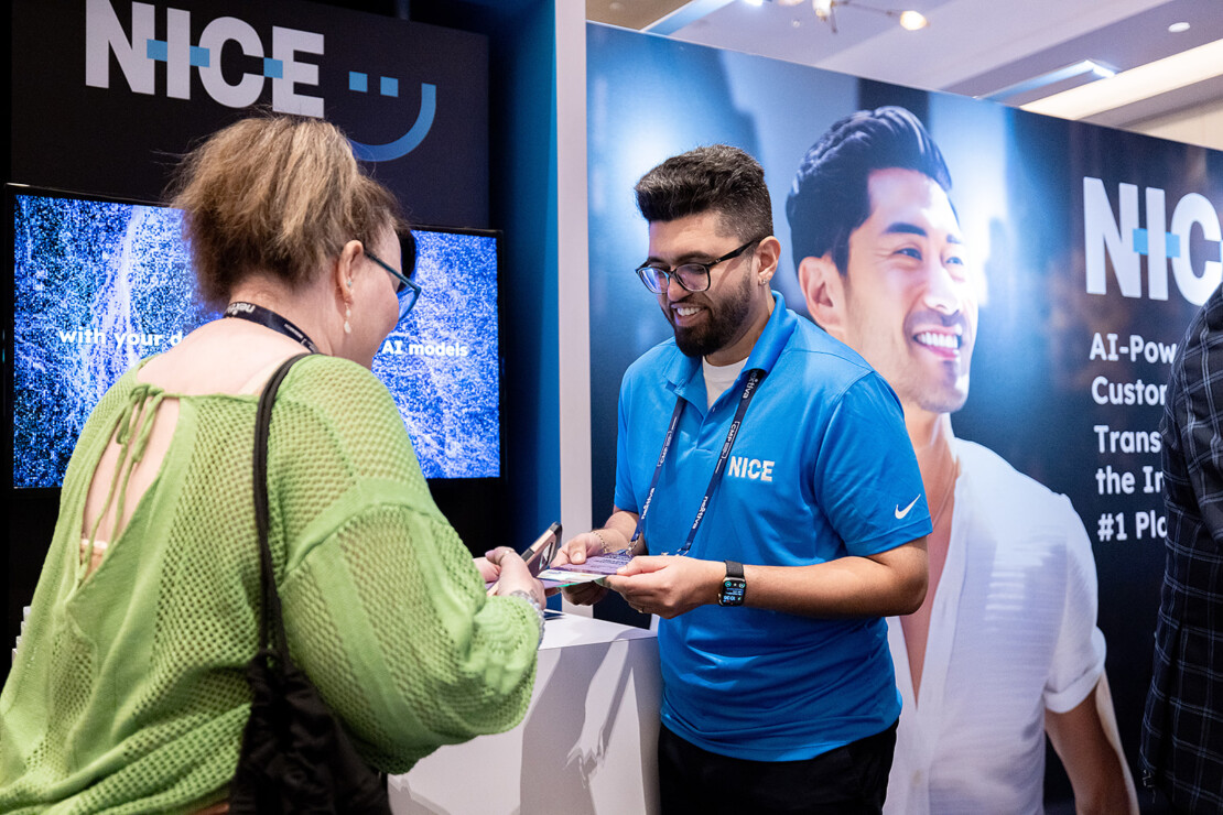 A man in a blue NICE shirt speaks with a woman at a trade show booth featuring NICE branding and promotional displays.