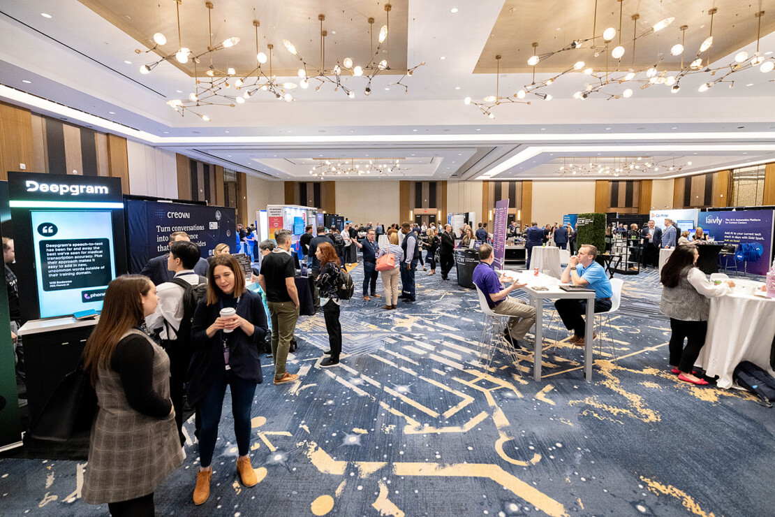 People networking and visiting booths at a tech conference in a large, well-lit room with modern decor and digital displays.