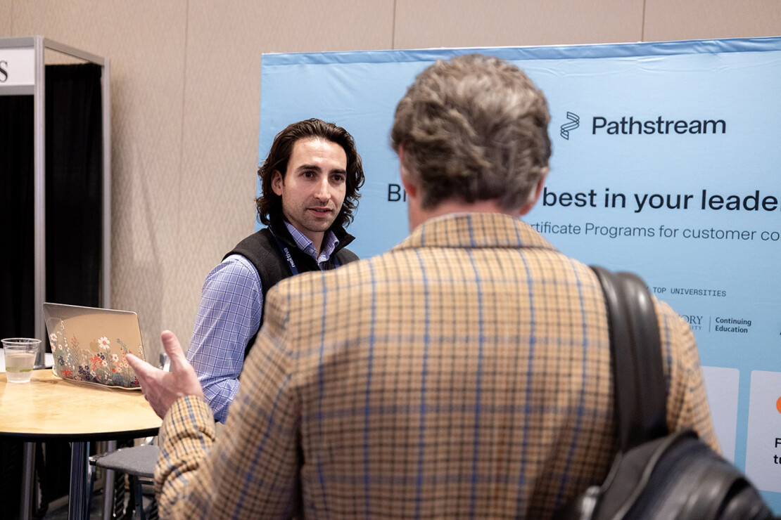 Two men have a conversation at a Pathstream booth; one faces the camera, while the other stands with his back turned, holding a bag.