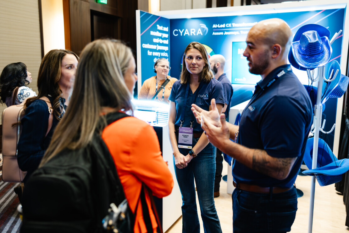 A group of people stand and talk at a tech conference booth with a "CYARA" display in the background.