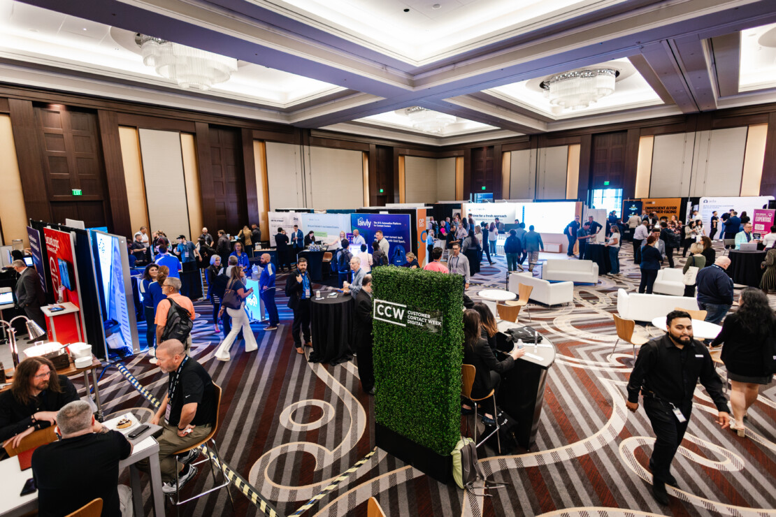 People interact at various booths and tables in a large, well-lit conference hall during a business convention or trade show.
