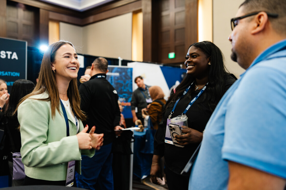 Three people stand and converse at a professional event, wearing name badges; one holds a drink cup, and others in the background interact near booths.