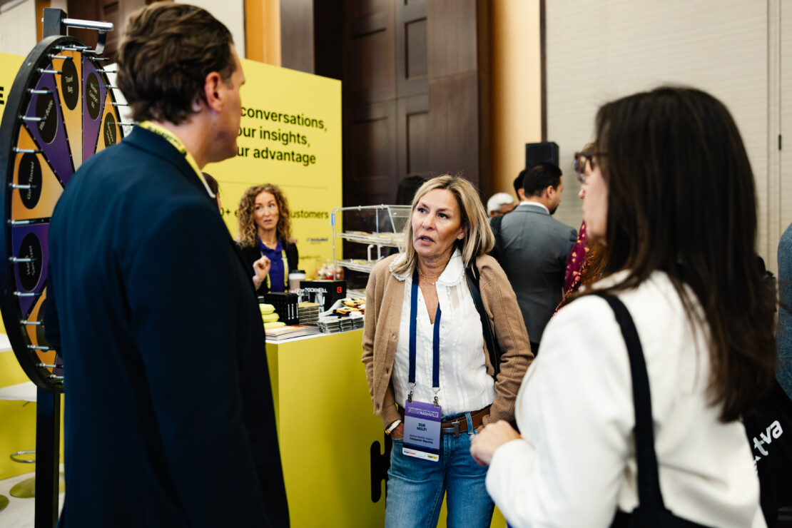 Three people stand talking near a yellow booth display with a prize wheel at a professional event or conference.