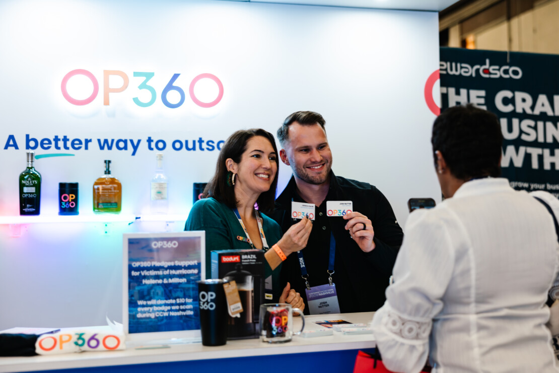 Two people at an OP360 booth smile and hold up business cards while talking to a third person at a business event.