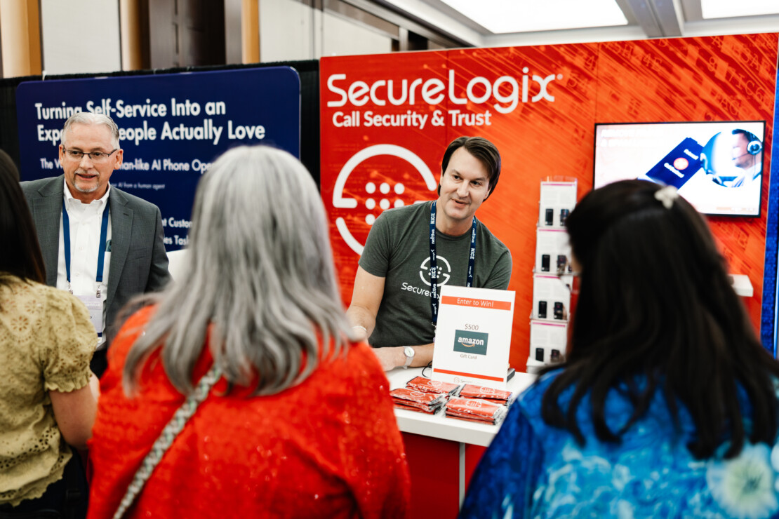 A man stands at a SecureLogix booth, talking to three people while promoting cybersecurity products at a conference.