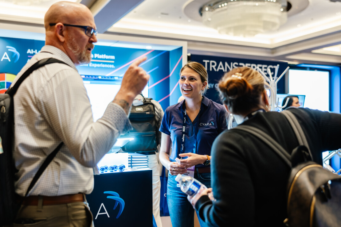 Three people interact and smile at a trade show booth, with technology displays and promotional materials visible in the background.
