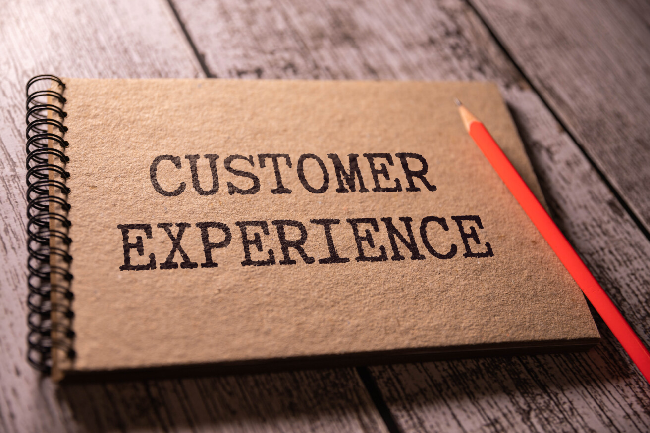 A spiral notebook with "Customer Experience" written on the cover, placed next to a red pencil on a wooden surface.