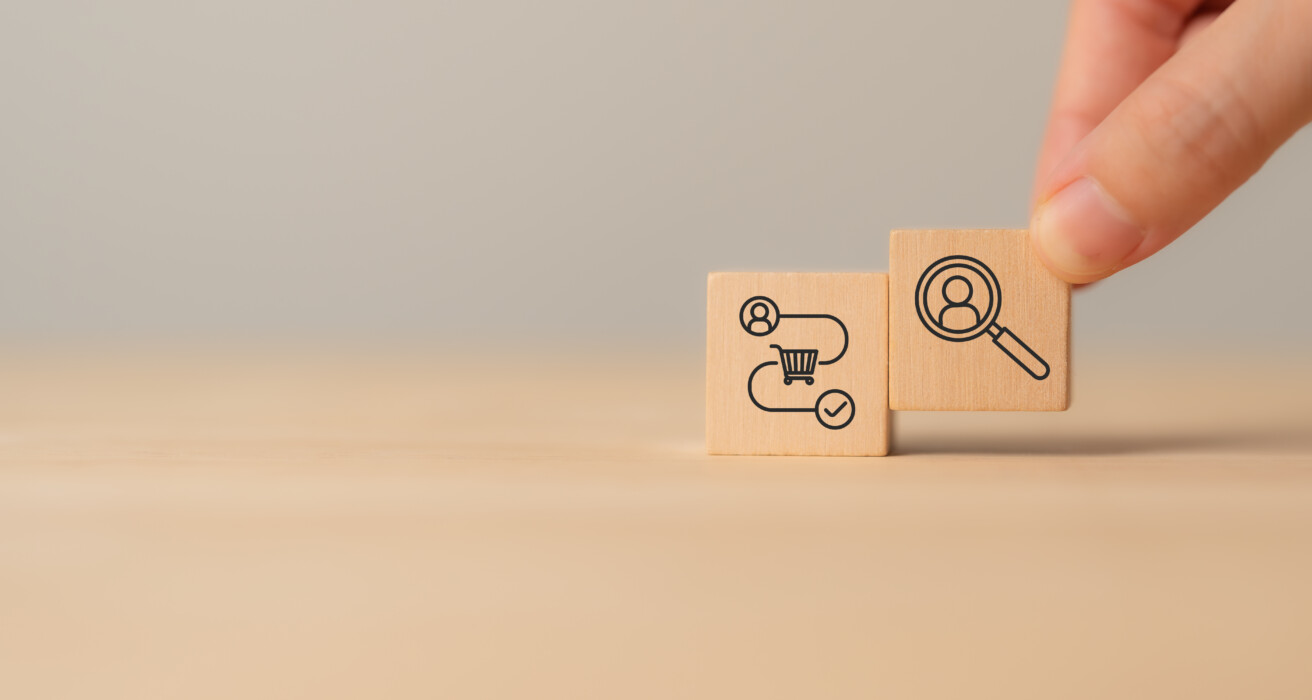 A hand holding a wooden block with a magnifying glass and person icon, next to a block with workflow and gear symbols on a plain background.