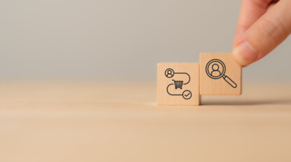 A hand holding a wooden block with a magnifying glass and person icon, next to a block with workflow and gear symbols on a plain background.