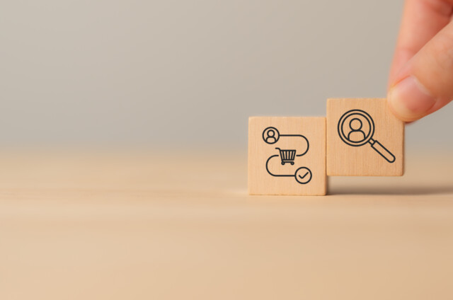 A hand holding a wooden block with a magnifying glass and person icon, next to a block with workflow and gear symbols on a plain background.