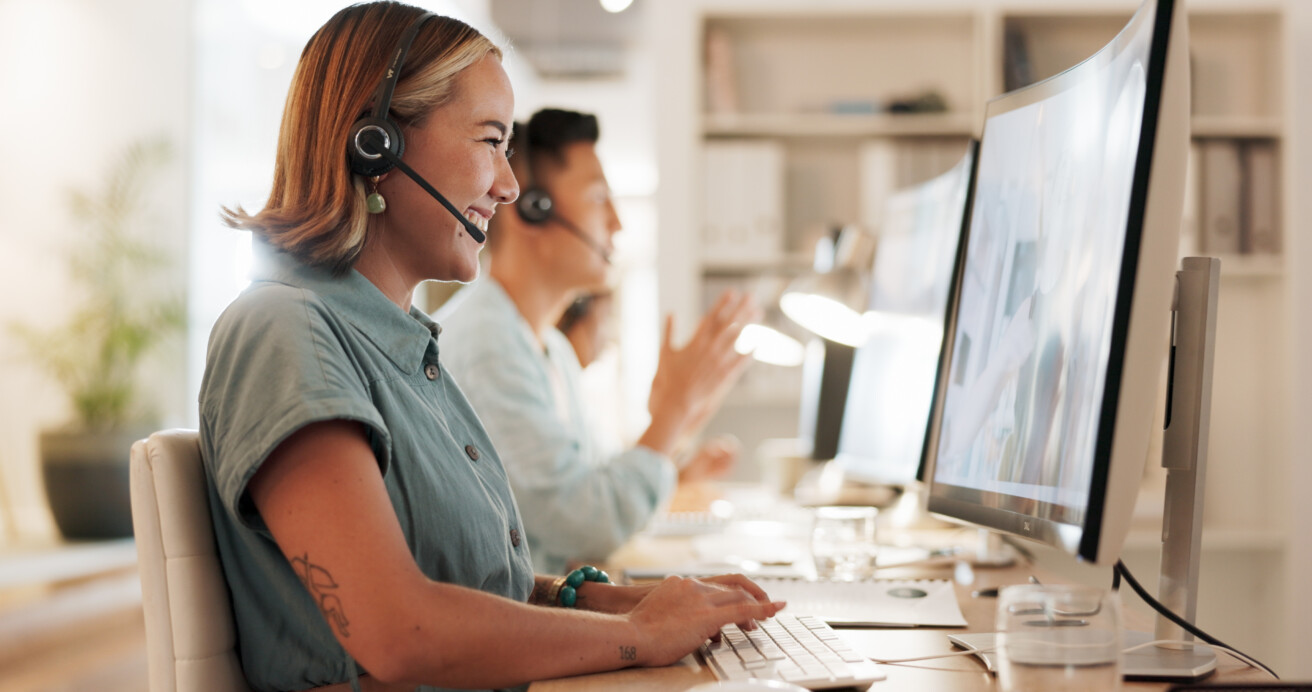 Two customer service representatives wearing headsets sit at desks, working on computers and smiling in a well-lit office environment.
