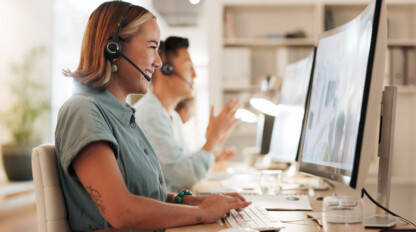 Two customer service representatives wearing headsets sit at desks, working on computers and smiling in a well-lit office environment.