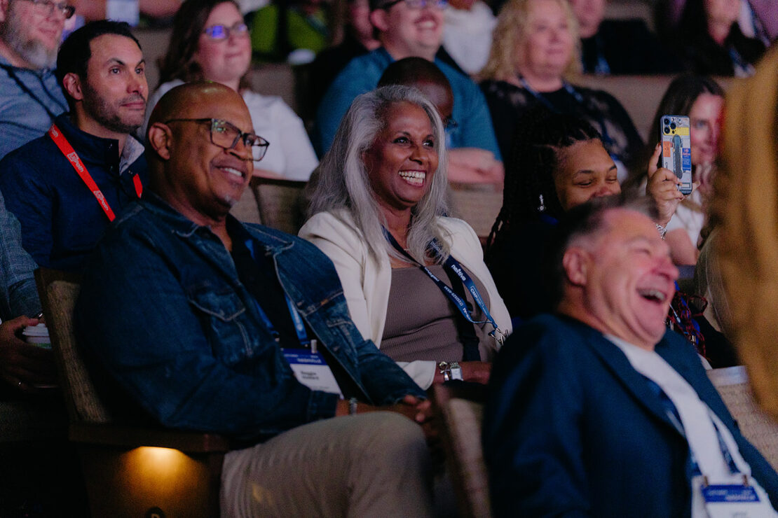 A group of people sit in an auditorium, smiling and laughing while watching an event or presentation.