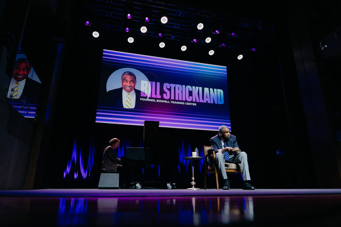 A man sits on stage beside a pianist, with a large screen behind them displaying "BILL STRICKLAND, FOUNDER, BIDWELL TRAINING CENTER" and a photo of Bill Strickland.