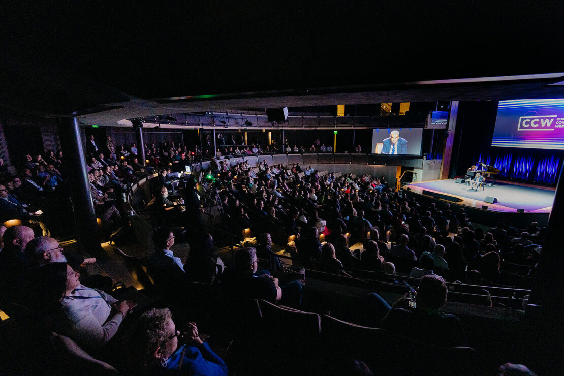 A large audience sits in a dimly lit auditorium, watching a speaker on stage who is also shown on a screen above.