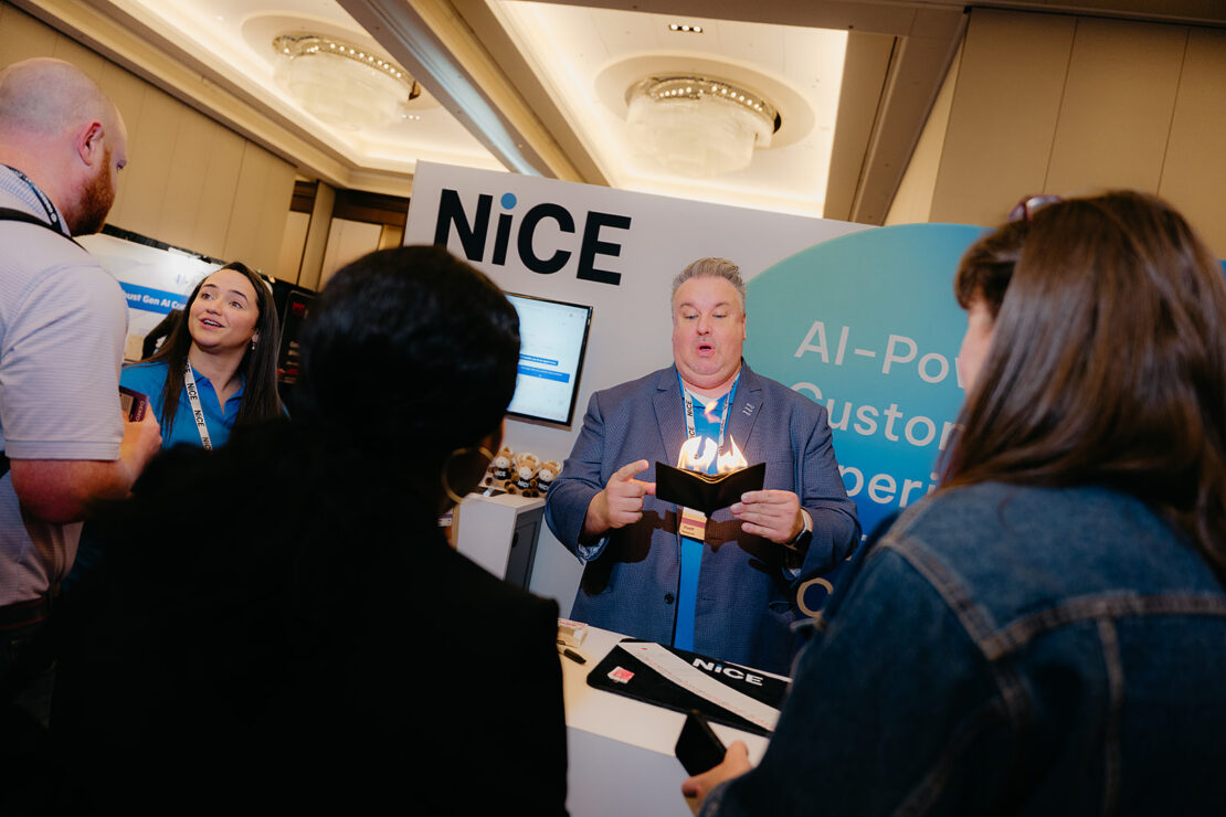 A man stands at a NiCE booth demonstrating a product to a small group of people at a conference or trade show.