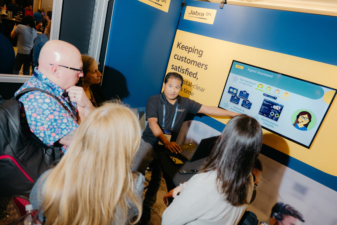 A man gives a presentation to a group of people at a booth, pointing at a display screen with information about an agent assistant.