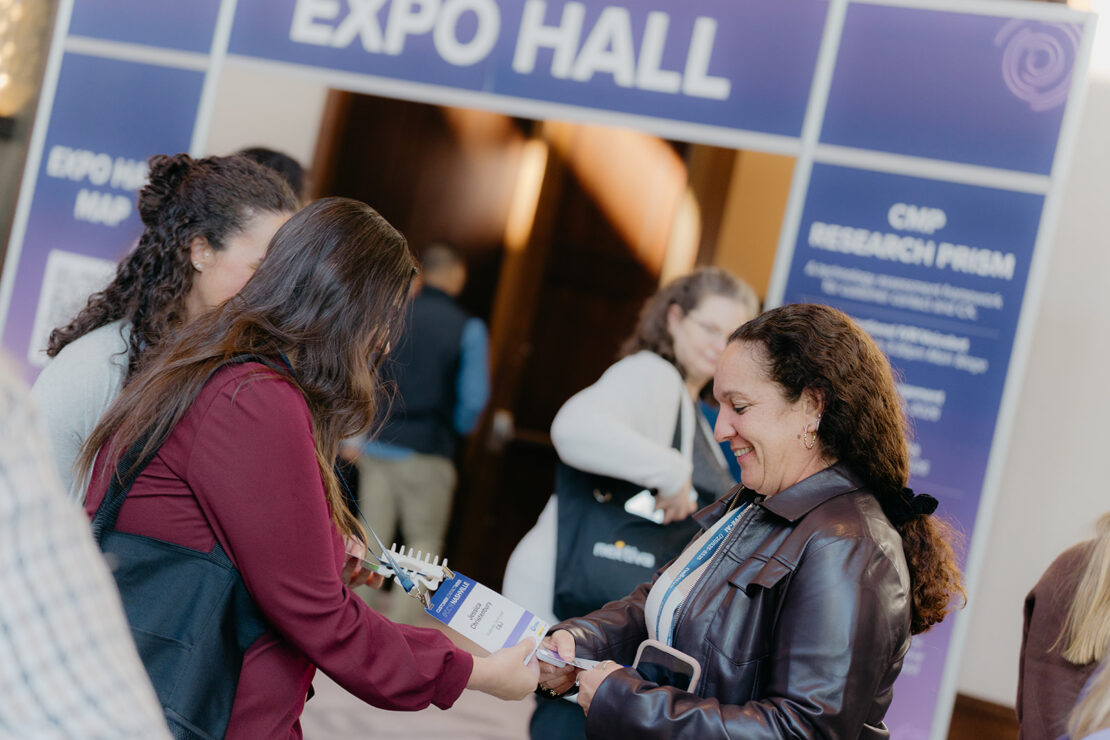 People interact and exchange materials near the entrance of an expo hall, with a large sign reading "EXPO HALL" visible in the background.