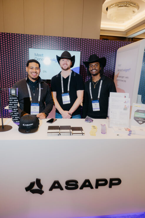 Three people wearing black hats stand behind an ASAPP booth at a conference, with promotional materials and a display screen in the background.