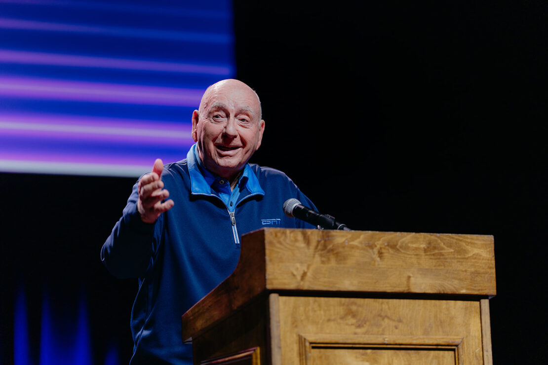 A man stands at a wooden podium, gesturing with one hand while speaking into a microphone, in front of a dark background with blue and purple light.