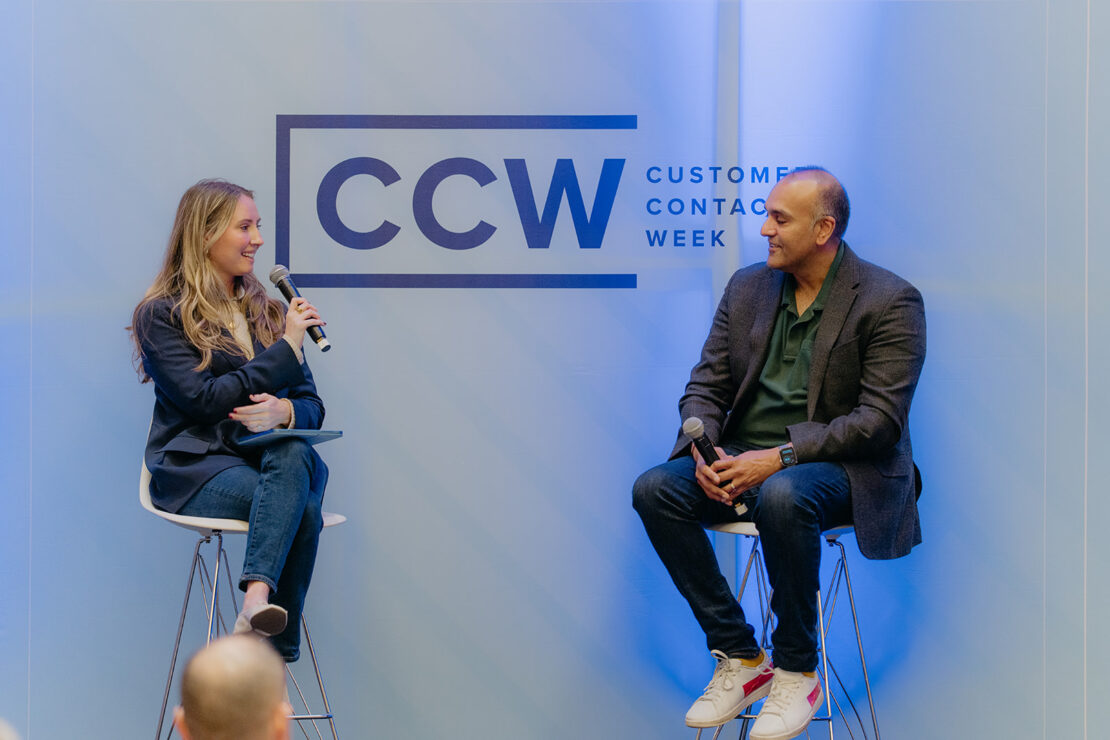 A woman and a man sit on stools holding microphones, engaged in conversation onstage at Customer Contact Week, with a CCW event sign in the background.